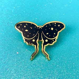 May include: Gold-tone enamel pin featuring a black butterfly with a galaxy design. The butterfly has a detailed design with stars and planets.
