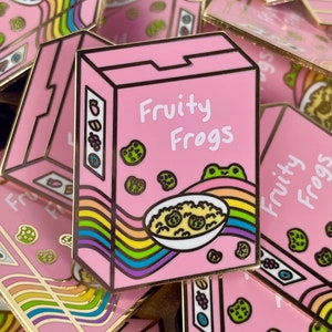May include: A pink enamel pin shaped like a cereal box with the text "Fruity Frogs" and a rainbow design. The box contains a bowl of cereal with green frog-shaped cereal pieces.