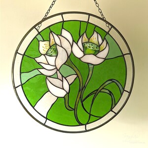 Elegant round stained glass panel featuring White lilies and a variegated green background, centered against a light wall, highlighting the detailed flower suncatcher design and the quality of the glasswork.