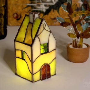 Può includere: Un portacandele a forma di casa in vetro colorato con pannelli di vetro giallo, verde e ambra. La casa ha un camino e una piccola porta. Un albero decorativo di vetro in un piccolo vaso è accanto alla casa.