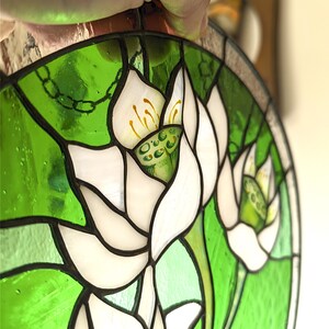Overhead close-up of the stained glass panel with White lilies, held by hand, highlighting the intricate Tiffany glass art soldering and the pearlescent texture of the white glass petals against the bright green background.