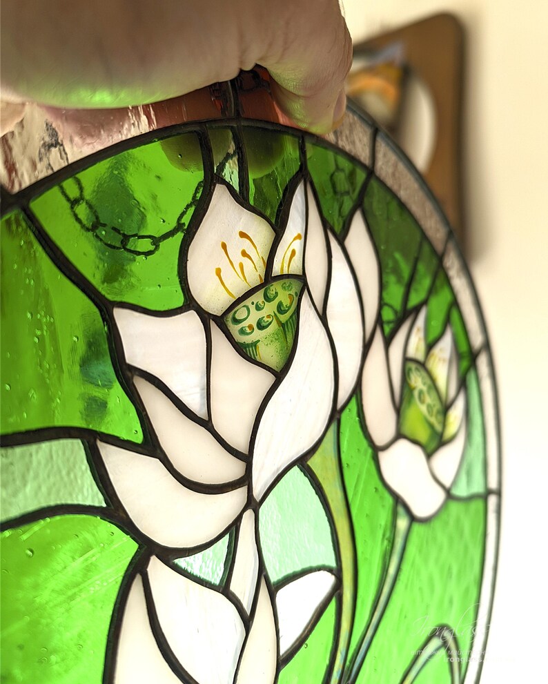 Overhead close-up of the stained glass panel with White lilies, held by hand, highlighting the intricate Tiffany glass art soldering and the pearlescent texture of the white glass petals against the bright green background.