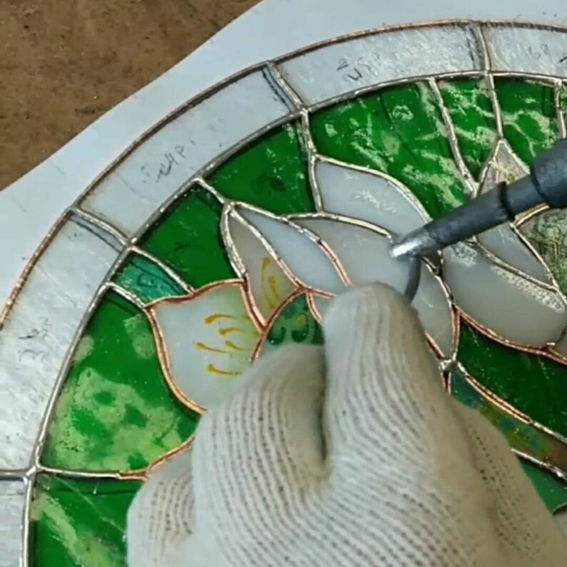 Close-up shot of the Tiffany glass art soldering process, showing a gloved hand skillfully fusing the copper-foiled White Lilies glass pieces with molten solder, demonstrating the meticulous handmade craftsmanship of the stained glass panel.