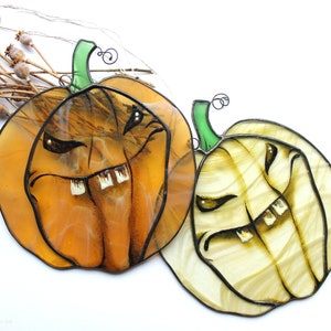 May include: Two stained glass pumpkins with spooky faces. The pumpkin on the left is orange with a toothy grin and the pumpkin on the right is white with a toothy grin.