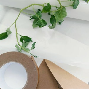 May include: A roll of brown packing tape, a brown envelope, and a white sheet of paper are arranged on a white surface. Green leafy vines are draped across the scene. The tape roll is partially unrolled, revealing the adhesive side.