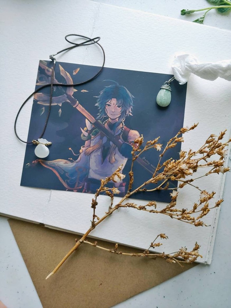 May include: An illustration of a character with blue hair and a spear, with a necklace featuring a white pendant and a green gemstone pendant. A dried plant stem and a white paper are also in the image.