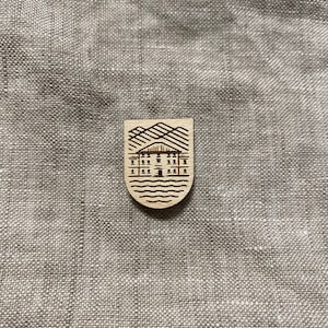 Może przedstawiać: Drewniana szpilka z czarno-białym rysunkiem domu z górami w tle. Dom ma drzwi i okna. Szpilka znajduje się na jasnobrązowym tle z tkaniny.