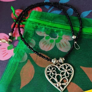 May include: A necklace featuring a strand of small black beads and a silver-toned heart-shaped pendant. The pendant has a cut-out floral design and is attached to the beaded chain with two decorative silver-toned metal beads on either side.