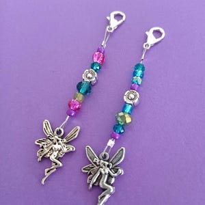 May include: Two silver-toned metal fairy charms attached to lobster claw clasps. Each charm features a series of colorful beads, including purple, pink, teal, and green faceted glass beads, along with small silver-toned flower-shaped spacers.