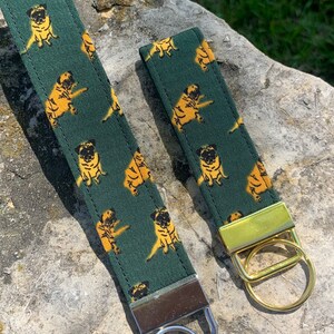 Puede incluir: Dos llaveros de tela verde con un patrón repetido de perros pug amarillos. El llavero de la izquierda tiene un anillo de llave plateado y el llavero de la derecha tiene un anillo de llave dorado.
