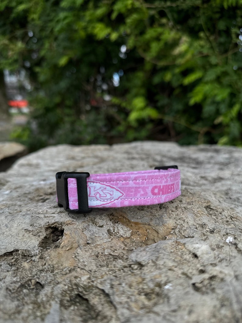Puede incluir: Collar de perro rosa con hebilla negra y logotipo blanco de los Kansas City Chiefs. El collar tiene un dise&ntilde;o estampado rosa y blanco.