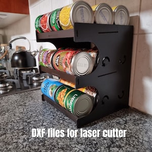 Puede incluir: Una estantería negra de tres niveles para guardar latas. La estantería está hecha de madera y tiene un diseño sencillo. Se muestra con varios productos enlatados en cada estante.