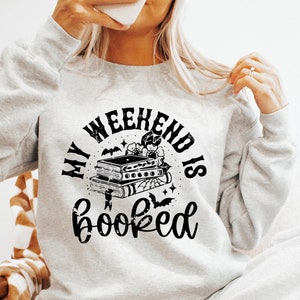 K&ouml;nnte beinhalten: Ein graues Sweatshirt mit einer schwarzen Grafik, auf der "My Weekend is Booked" steht, mit einem Stapel B&uuml;cher, Flederm&auml;usen und Sternen. Die Grafik ist in einem Kreis.