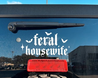 May include: A black car with a white vinyl sticker that reads "feral housewife" with bat silhouettes. The sticker is on the back window of the car. The car has a red brake light and a spare tire.