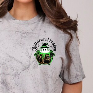 May include: A gray tie-dye t-shirt with a black and green graphic of a ghost in a witch's hat inside a cauldron. The text "You are not broken" is written in a black gothic font around the graphic.