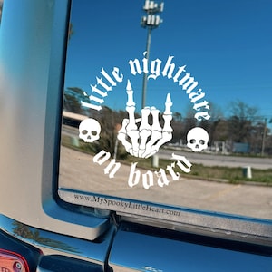 May include: A white vinyl sticker on a black car. The sticker says "little nightmare on board" in a gothic font with a skeleton hand giving the middle finger and two skulls on either side. The website www.myspookylittleheart.com is printed at the bottom.