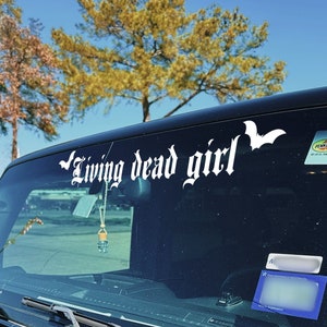 May include: A black Jeep Wrangler with a windshield sticker that reads "Living dead girl" with a bat symbol. The Jeep is parked in a parking lot with trees in the background.