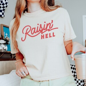 May include: A woman wearing a light yellow cropped t-shirt with the text "Raisin' Hell" in pink. She is wearing light green shorts and holding a cup of coffee.