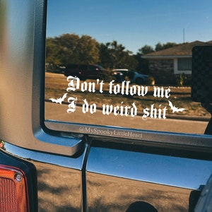 May include: A black car with a white vinyl sticker on the back window that reads "Don't follow me I do weird shit" with a bat graphic on either side of the text. The sticker also includes the text "@MySpookyLittleHeart".
