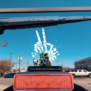 May include: A white skeleton hand making a peace sign with flowers and a smiley face. The sticker is on the back window of a black car. The text on the sticker reads "www.MySpookyLittleHeart.com".