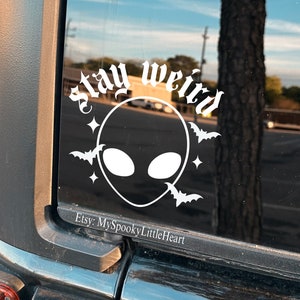May include: A black car with a white vinyl sticker on the back window. The sticker says "Stay Weird" in a gothic font and features an alien face with bats flying around it. The sticker also includes the text "Etsy: MySpookyLittleHeart".