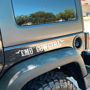 May include: Black Jeep Wrangler with a white vinyl decal that reads "EMO COWGIRL" with bat wings on either side of the text. The decal is on the back passenger side of the vehicle.