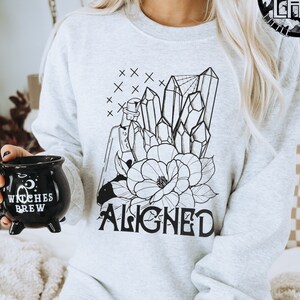 May include: A gray sweatshirt with a black and white graphic of crystals, a flower, and a potion bottle. The text "ALIGNED" is printed below the graphic.