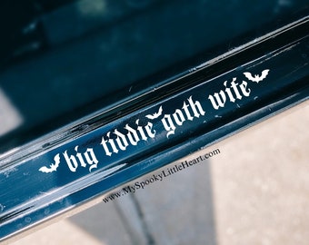 May include: A black car door with a white vinyl decal that reads "big tiddie goth wife" with bat silhouettes on either side. The decal also includes the website "www.MySpookyLittleHeart.com".