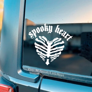 May include: A white vinyl sticker with a spooky heart design featuring a skeleton ribcage. The sticker is on the back window of a black car. The text "spooky heart" is written in a gothic font around the heart. The text "Etsy: MySpookyLittleHeart" is written below the heart.