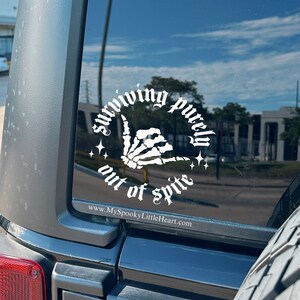 May include: A white vinyl sticker on a black car window. The sticker features a skeleton hand giving the shaka sign with the text "surviving purely out of spite" around the hand. The text "www.MySpookyLittleHeart.com" is below the sticker.