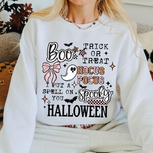 May include: White Halloween sweatshirt with the text "Boo Trick or Treat Hocus Pocus Spooky Halloween". The design includes a ghost, bow, and bat graphics. The sweatshirt is part of a Halloween-themed collection.