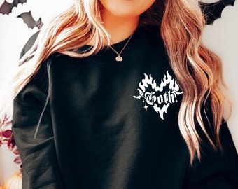 May include: A black sweatshirt with a white graphic of a heart with flames and the word "goth" inside. The sweatshirt is being worn by a person sitting on a wooden surface.