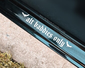 May include: A black car door with a white vinyl decal that reads "alt baddies only" with a bat on either side of the text. The decal is on a black car door with a black trim. The car is parked on a sidewalk next to a patch of green grass.