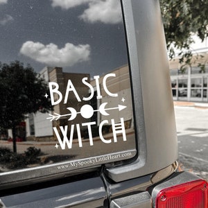 May include: A white vinyl decal on a black vehicle window. The decal reads "BASIC WITCH" in a stylized font, with an arrow, moon, and star design. The website address "MySpookyLittleHeart.com" is also visible.