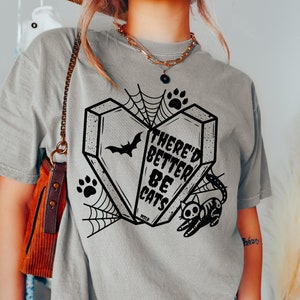 May include: A gray t-shirt with a black graphic of a coffin shaped like a heart with a bat, spider web, and paw prints. The text on the coffin reads "There'd Better Be Cats".