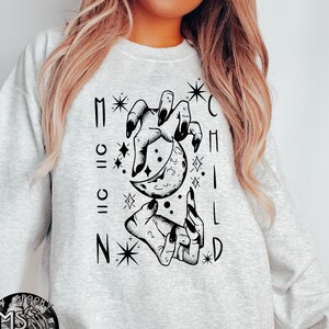 May include: A gray sweatshirt with a black graphic print of two hands holding a crescent moon. The text "MOON CHILD" is printed vertically around the moon.