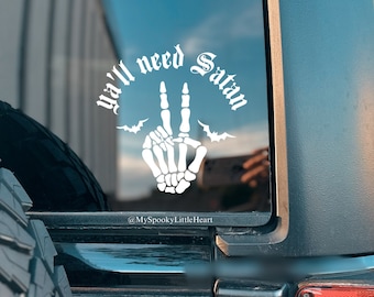 May include: A white vinyl sticker on a black car window. The sticker features a skeleton hand making a peace sign with the text "Ya'll need Satan" in a gothic font. The sticker also includes two bats on either side of the hand.