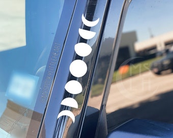 May include: A black car with a white moon phase decal on the driver's side window. The decal is a vertical line of moons in different phases, starting with a crescent moon and ending with a full moon. The decal is on the black trim of the window, and the car is parked in a parking lot.