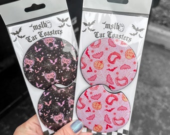 May include: Two sets of car coasters featuring a spooky Halloween theme. One set has a black background with bats, a spiderweb, and the word "Ouija" repeated. The other set has a pink background with bats, a pumpkin, and a red heart. Both sets are packaged in clear plastic bags with the brand name "mslk's Car Toasters" printed on the front.