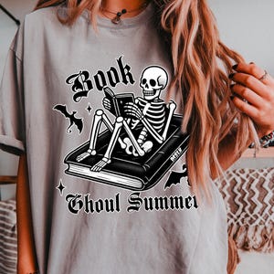 May include: Gray t-shirt featuring a skeleton reading a book with the words "Book Ghoul Summer" in a gothic font. The design includes bats and stars, creating a spooky summer theme.