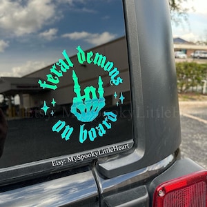 May include: A teal blue car decal with the text "Feral Demons on Board" in a gothic font. The decal features a skeleton hand giving the rock and roll hand gesture.