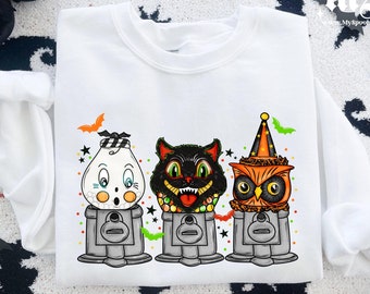 May include: White sweatshirt with a graphic of three Halloween characters in gumball machines. The characters are a ghost, a black cat, and an owl. The gumball machines are decorated with Halloween colors and symbols.