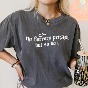 Op de afbeelding: Donkergrijs T-shirt met de tekst "the horrors persist but so do i" in witte gotische letters. Het shirt wordt gecombineerd met een gestreepte broek en een glas met luipaardprint en een houten deksel. Een neon pompoen staat op de achtergrond.