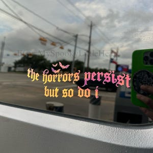 May include: A holographic vinyl decal applied to a window, featuring the text "the horrors persist but so do i" in a gothic-style font. The design includes three small bat silhouettes above the text, with a color-shifting gradient effect.