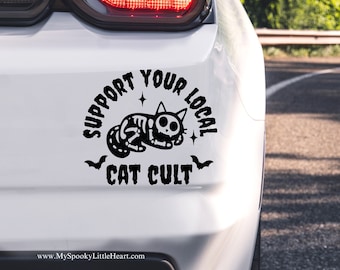 May include: A white car with a black and white sticker on the back bumper. The sticker says "Support Your Local Cat Cult" and features a cartoon skeleton cat.