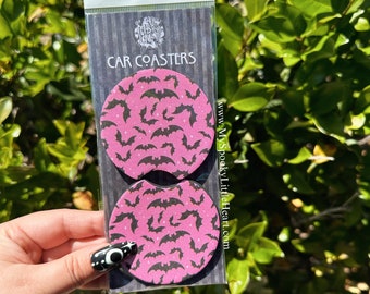 May include: Two pink round car coasters with a black bat pattern. The coasters are packaged in a clear plastic bag with a black and white striped backing card. The card reads "CAR COASTERS" and "www.MySpookyLittleHeart.com".