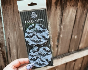May include: Two black and white car coasters with a bat pattern. The coasters are packaged in a clear plastic bag with a white header that says "CAR COASTERS" and a website address.