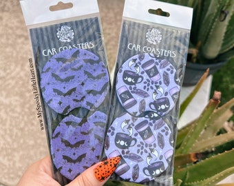 May include: Two sets of car coasters featuring a spooky Halloween theme. One set has a purple background with bats and the other has a purple background with coffee cups, ghosts, and crystals. The packaging says "Car Coasters".