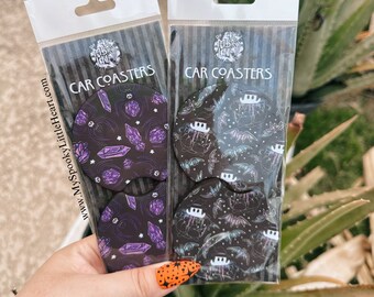 May include: Two sets of car coasters featuring a spooky, gothic theme. One set has a purple and black design with crystals and stars. The other set has a black and white design with bats and a crescent moon. The packaging says "CAR COASTERS" and "www.MySpookyLittleHeart.com".