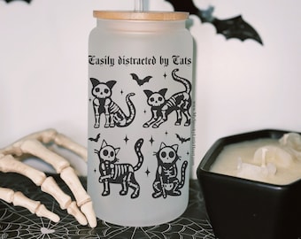 May include: A frosted glass tumbler with a bamboo lid and a straw. The tumbler has a black and white design of skeleton cats with the text "Easily distracted by Tats" and a website address. The tumbler is sitting on a black and white spiderweb pattern surface.